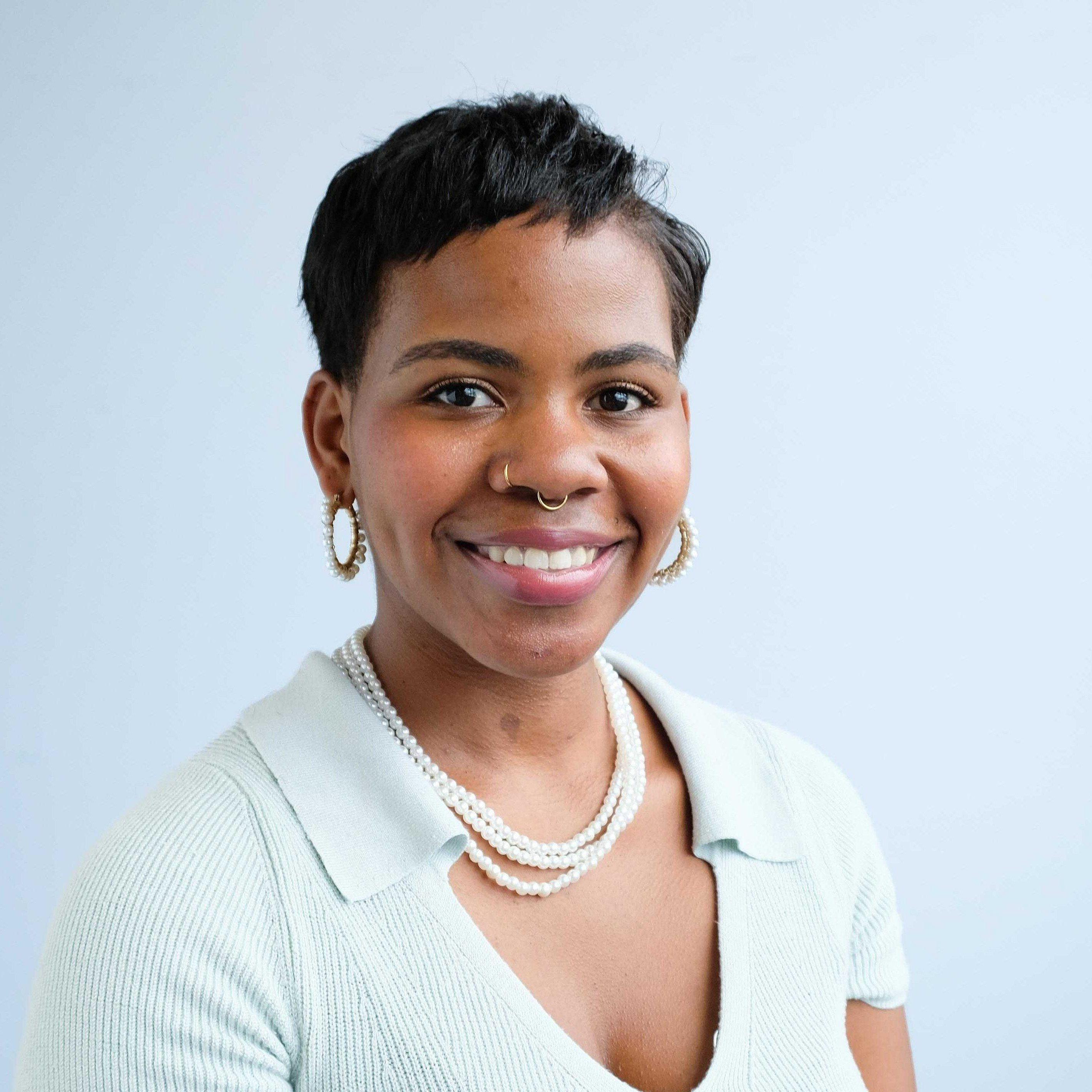 A woman with short black hair, wearing pearl earrings, a pearl necklace, and a light blue short-sleeve top, smiles while posing in front of a light blue background.